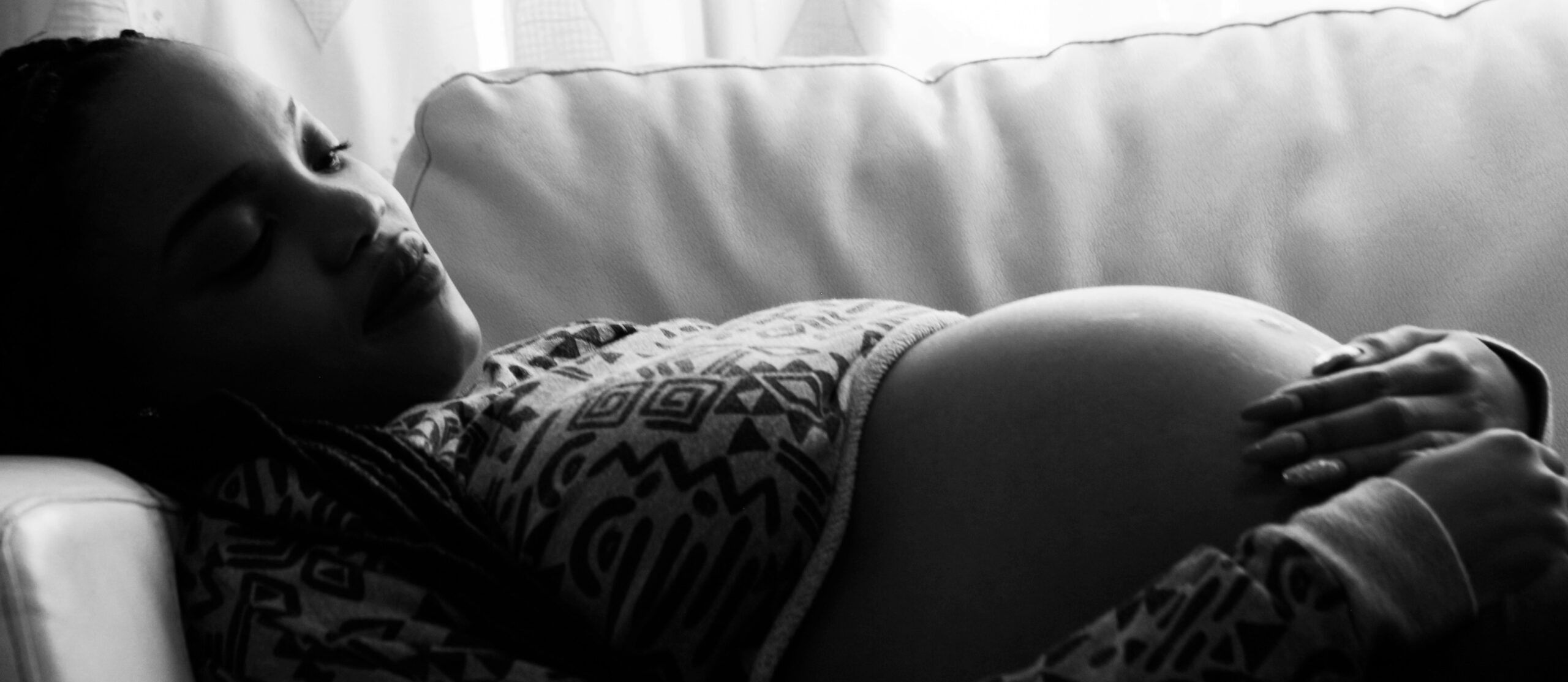 Schwangere Frau liegt mit geschlossenen Augen auf einem Sofa und hält beide Hände auf ihrem Bauch, Schwarz-Weiß-Fotografie