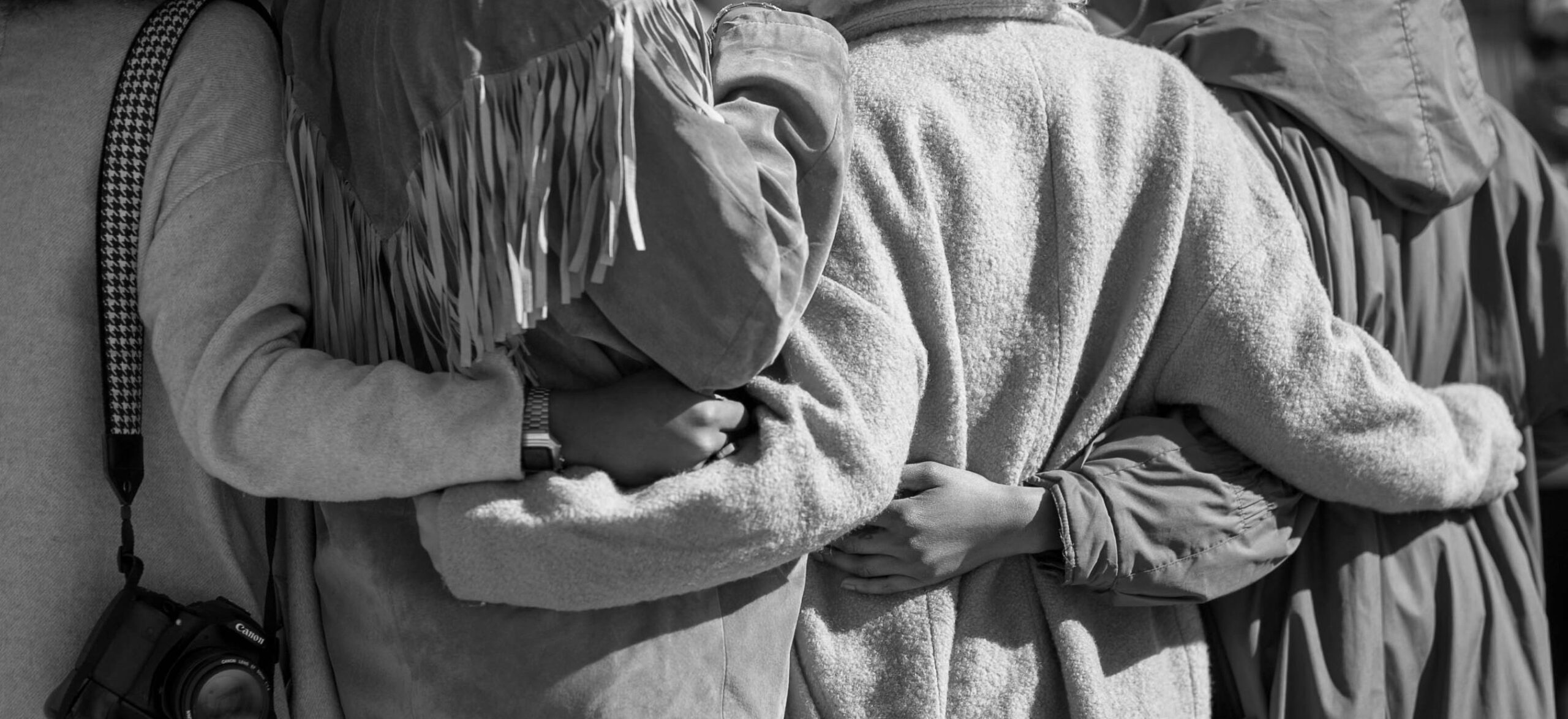 Personen von hinten fotografiert, Arme umeinander gelegt, eine trägt eine Fransenjacke, eine Kamera hängt an einem Trageriemen, Schwarz-Weiß-Fotografie