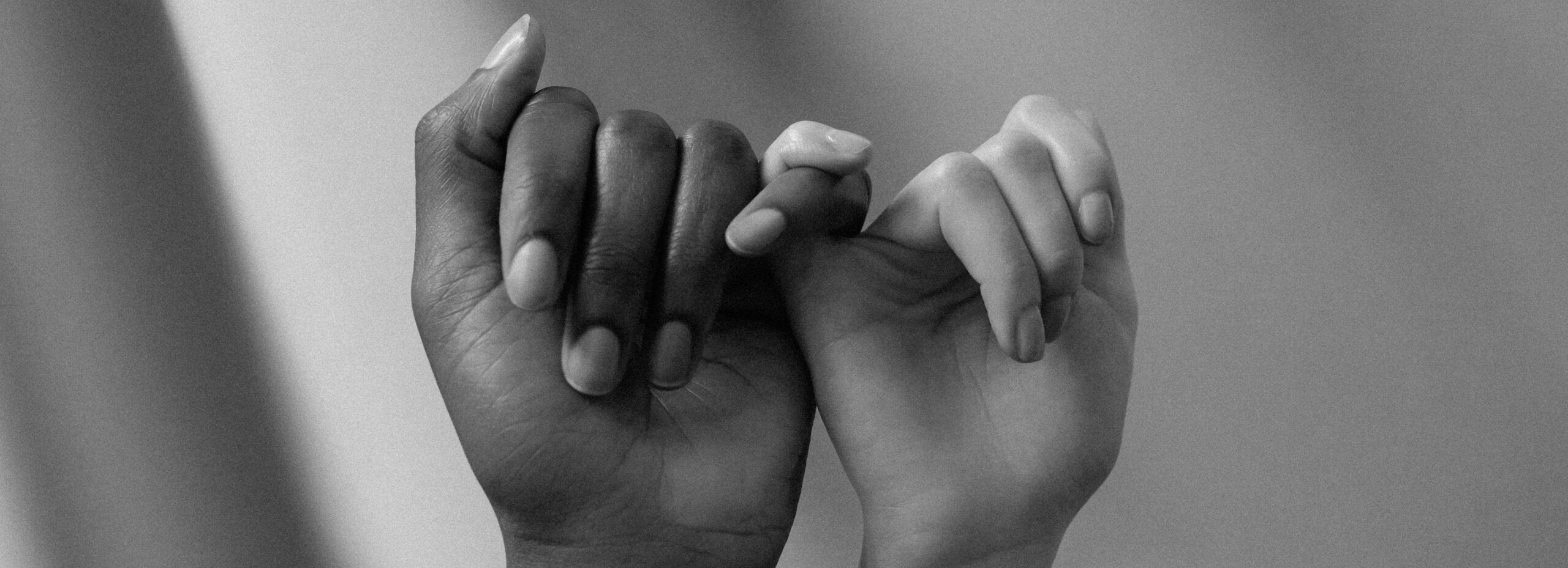 Zwei ineinander verschränkte Hände, Schwarz-Weiß-Aufnahme, mit dem kleinen Finger ineinandergehakt, vor unscharfem grauem Hintergrund.